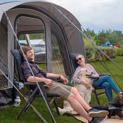 Vor einem Wohnmobilvorzelt sitzen zwei Personen auf schwarzen Dometic Opulence Firenze Relaxern, begleitet von zwei Hunden auf dem Campingplatz.