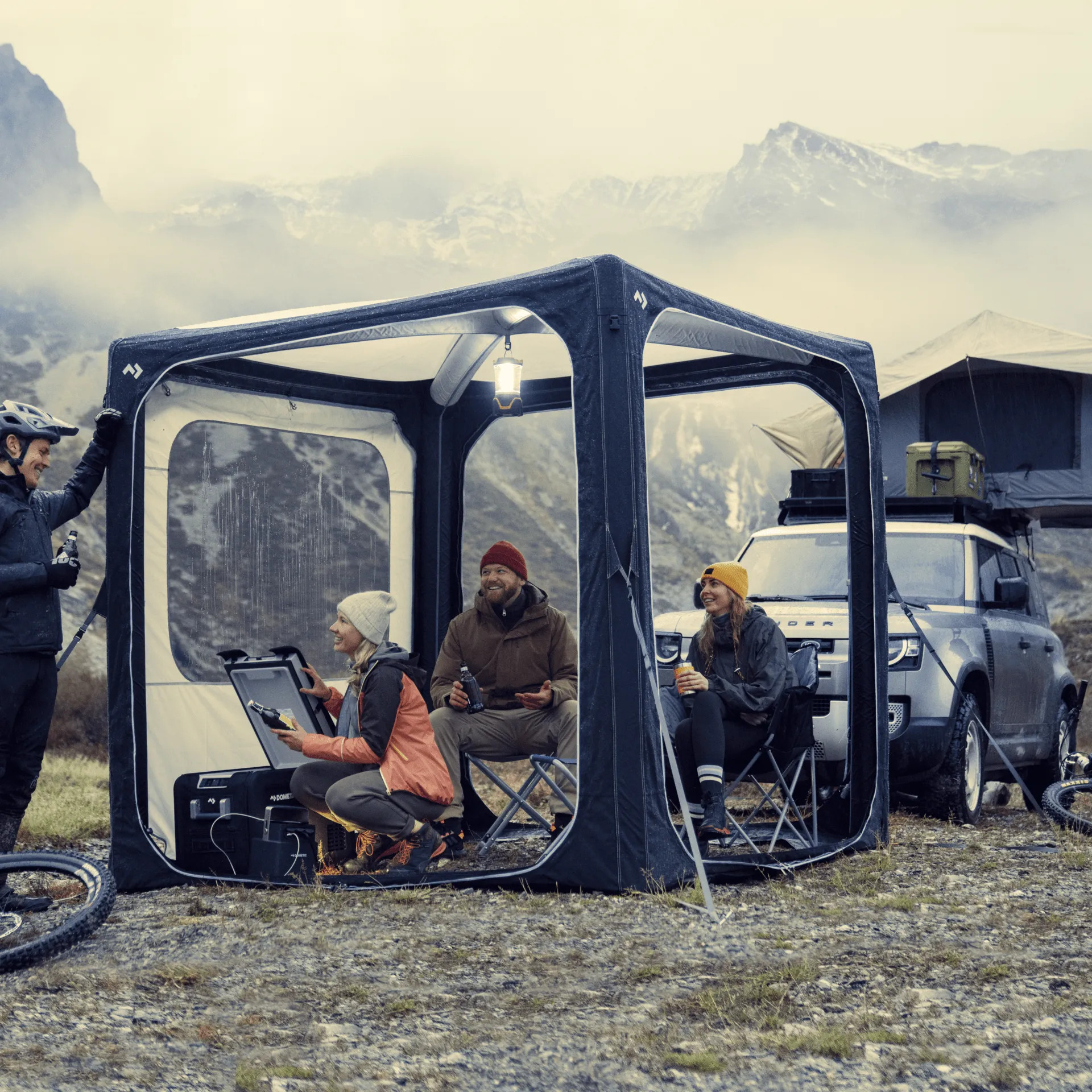 Gesellige Campingrunde im Dometic Hub Pavillon vor Bergkulisse mit Fahrzeug und Outdoor Ausruestung bei kuehler Witterung
