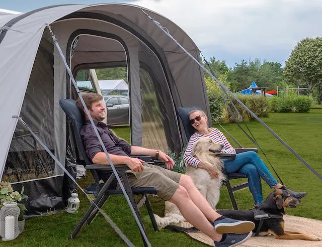 Vor einem Wohnmobilvorzelt sitzen zwei Personen auf schwarzen Dometic Opulence Firenze Relaxern, begleitet von zwei Hunden auf dem Campingplatz.