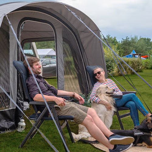 Vor einem Wohnmobilvorzelt sitzen zwei Personen auf schwarzen Dometic Opulence Firenze Relaxern, begleitet von zwei Hunden auf dem Campingplatz.
