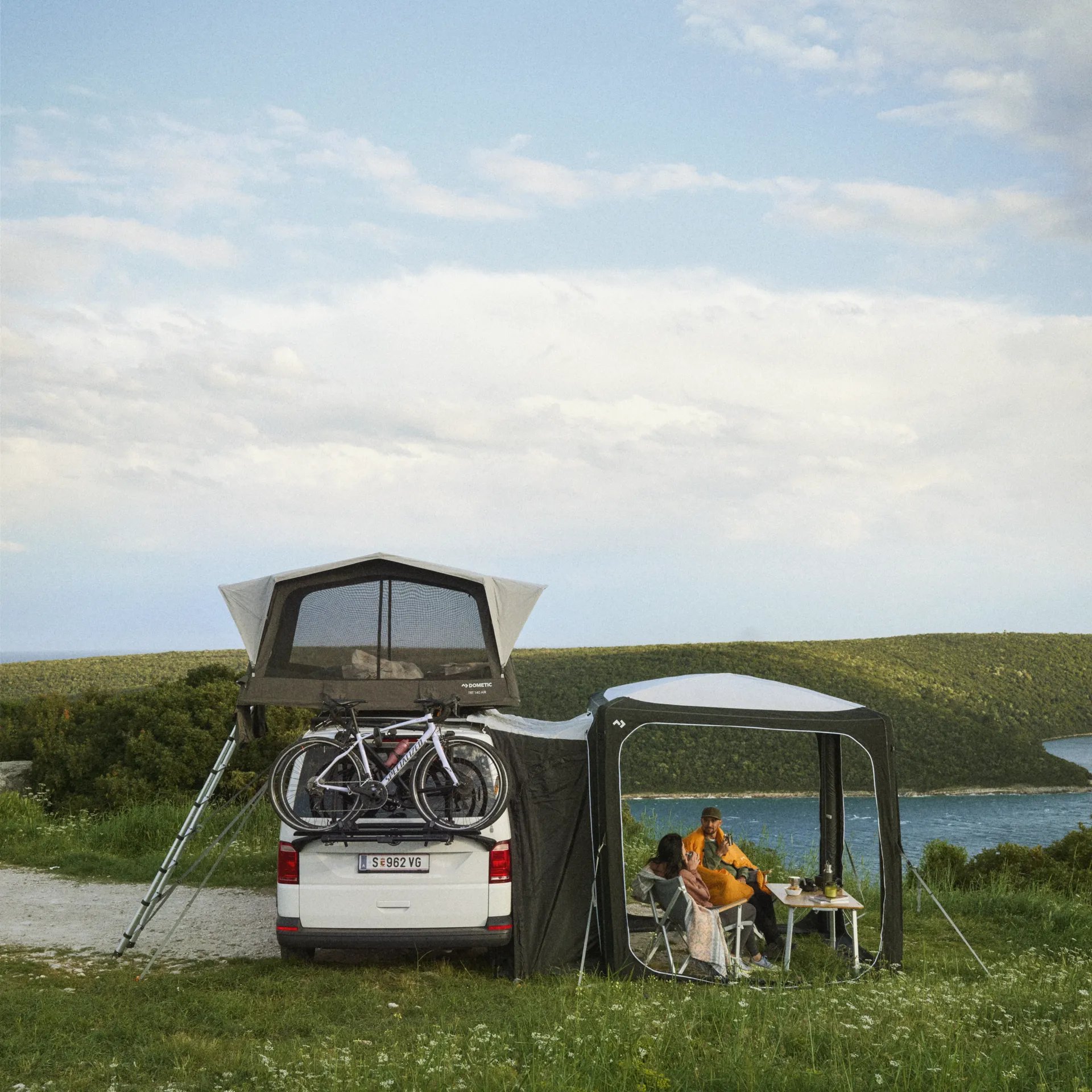 Idyllische Campszene mit Dometic Hub Pavillon neben Fahrzeug mit Dachzelt, Personen sitzen entspannt mit Blick auf Wasser und Landschaft