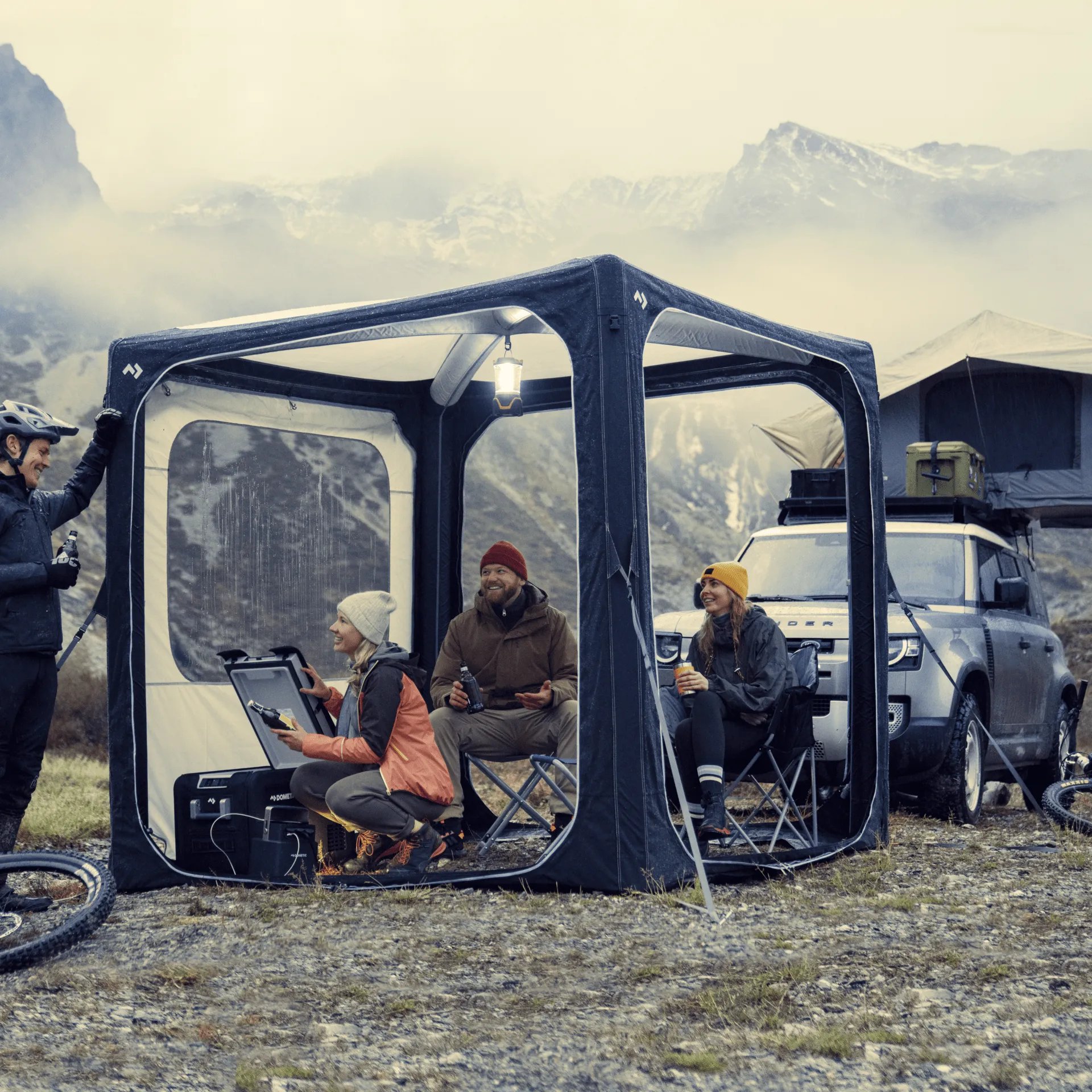 Gesellige Campingrunde im Dometic Hub Pavillon vor Bergkulisse mit Fahrzeug und Outdoor Ausruestung bei kuehler Witterung