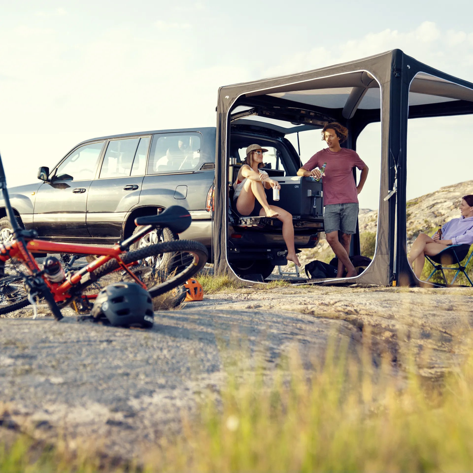 Campingalltag mit Dometic Hub Pavillon am Fahrzeug, Personen entspannen im Schatten neben Fahrrad und Outdoor Ausruestung