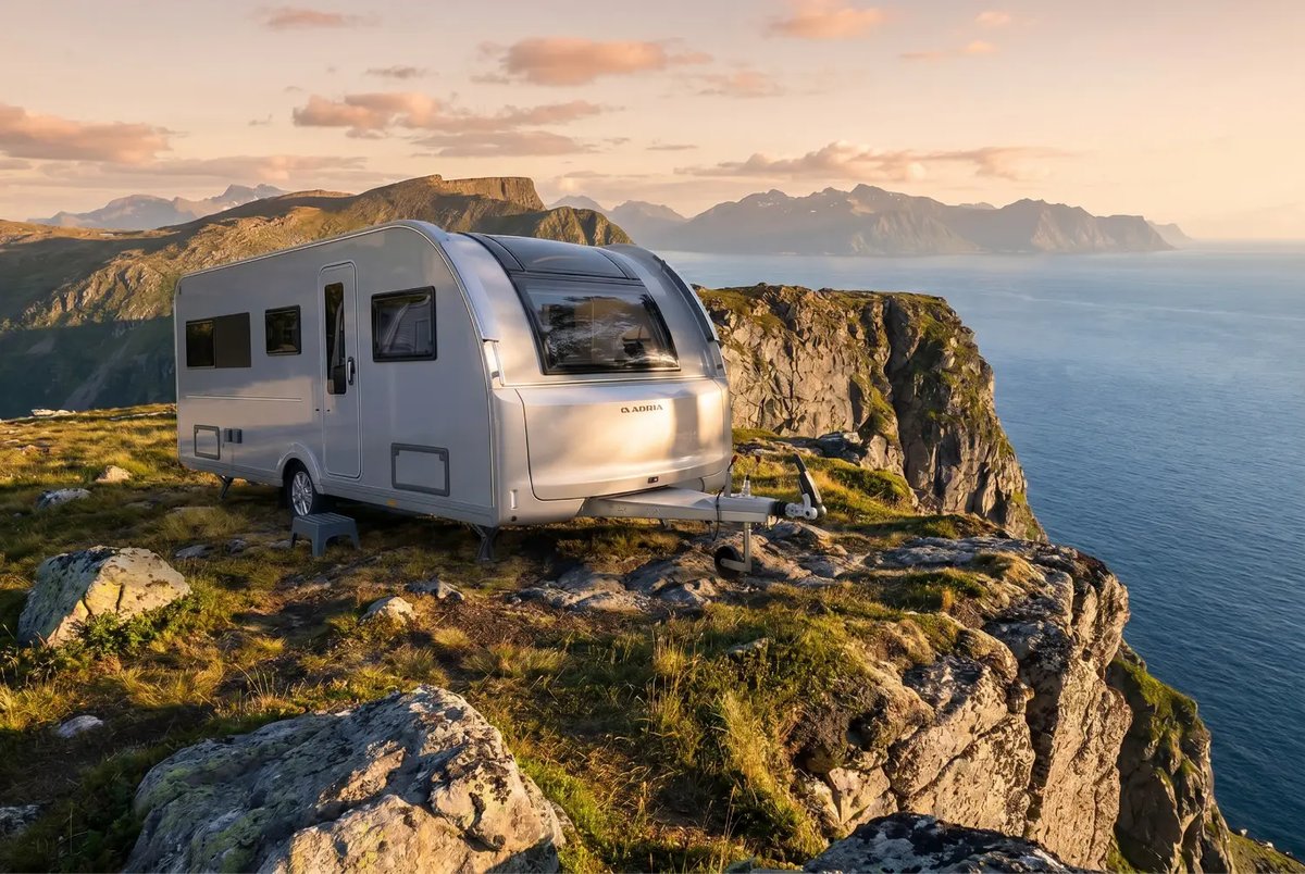 Der neue silberne Adria Alpina Wohnwagen auf einem Felsplateau hoch über dem Meer, im Hintergrund eine Bergkette