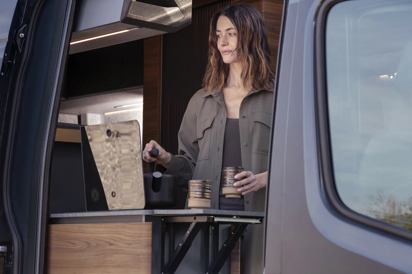 Adria Supertwin 2026 - Frau an der geöffneten Schiebetür bereitet Kaffee auf der seitlichen Arbeitsplattenerweiterung zu