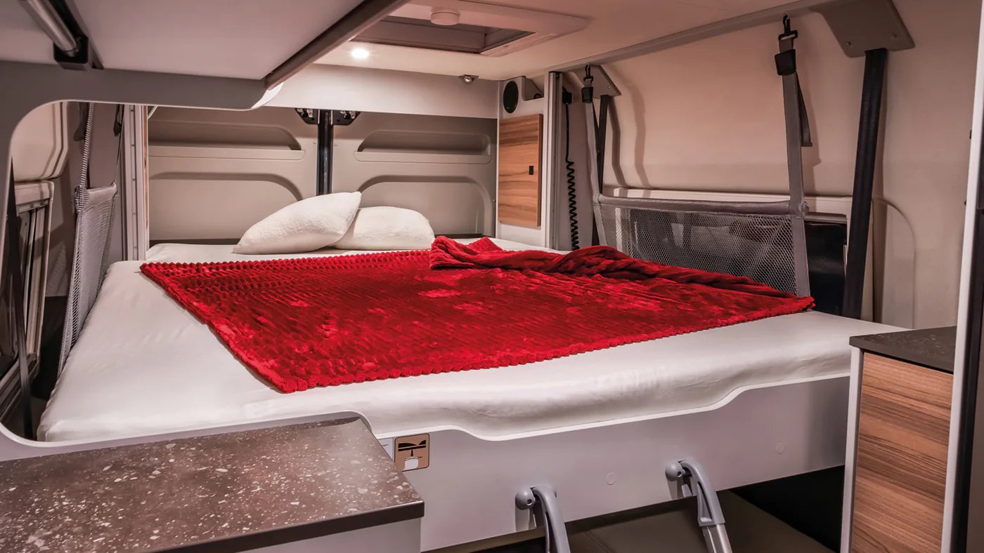 Abgesenktes Hubbett über der Hecksitzgruppe im Bürstner B66 Camper Van mit roter Decke, Haltegurten und Sicherungsnetz