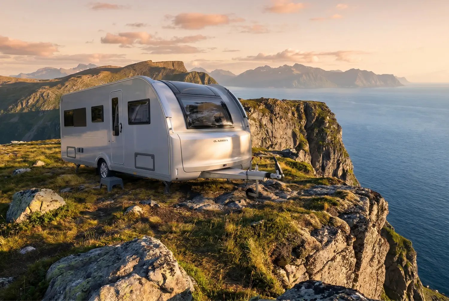 Der neue silberne Adria Alpina Wohnwagen auf einem Felsplateau hoch über dem Meer, im Hintergrund eine Bergkette