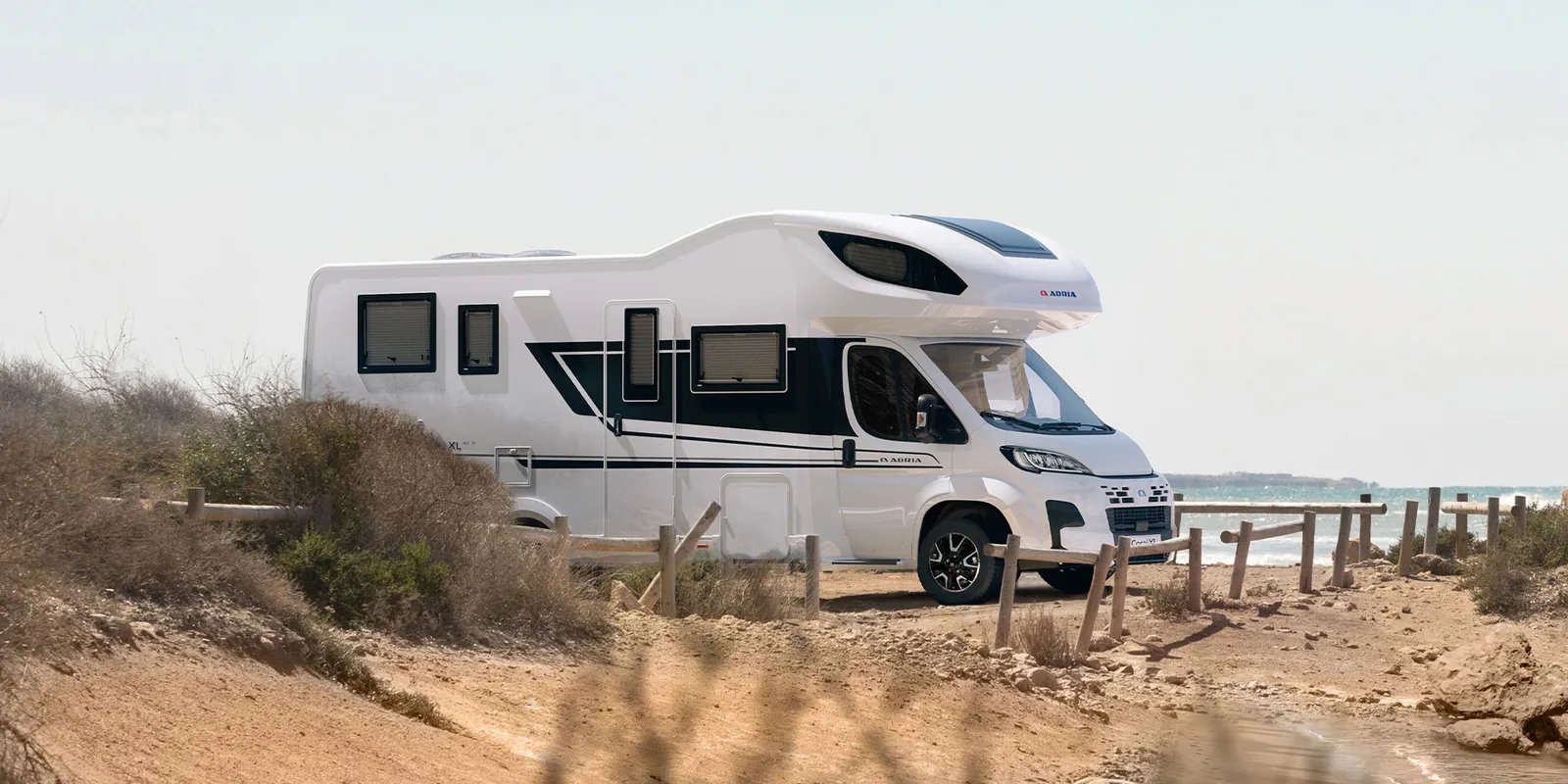 Adria Coral XL 2026 auf Fiat-Basis an einem Sandweg mit Holzzaun, Meer im Hintergrund