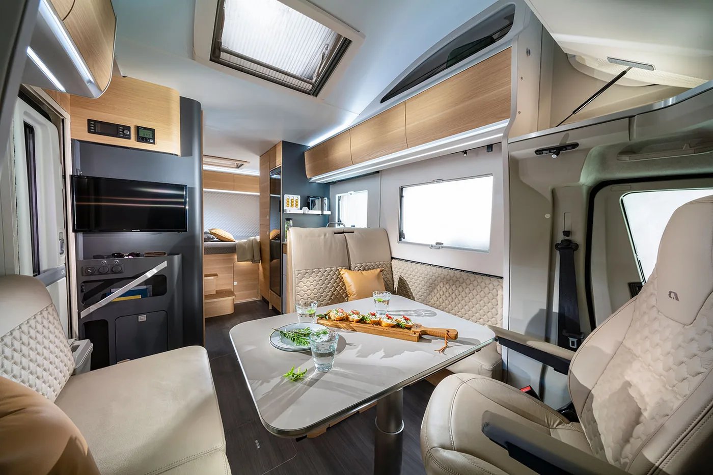 Adria Coral XL 2026 - Wohnraum-Gesamtblick mit Dinette, Multimedia-Wand und Alkoven über der Fahrerkabine