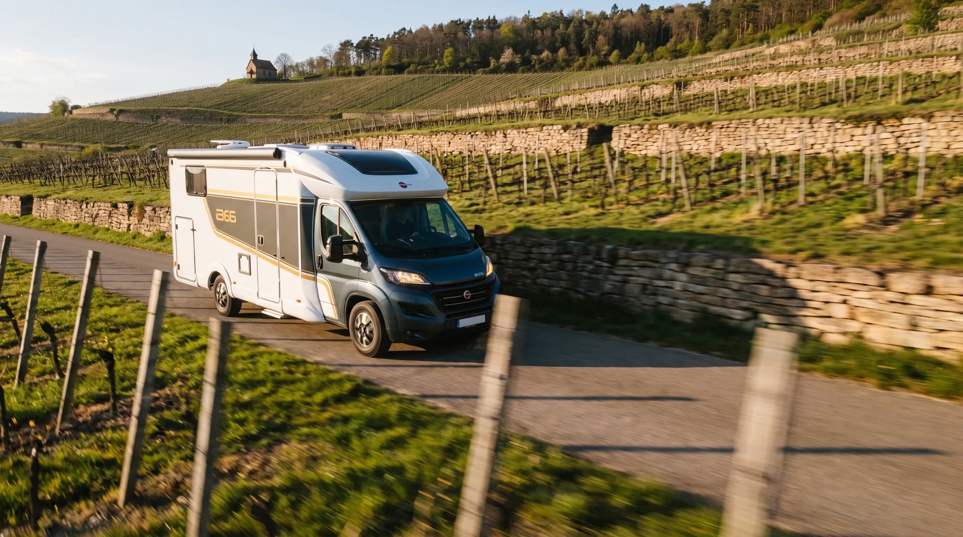 Bürstner B66 Wohnmobil auf einer schmalen Straße durch Weinbergterrassen, auf der Kuppe dahinter eine kleine Kapelle
