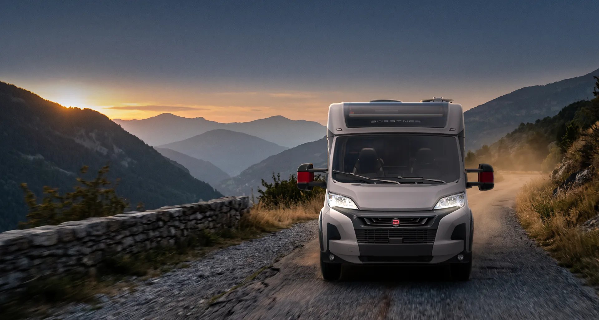 Weisses Bürstner Reisemobil auf einer schmalen Bergstrasse bei Sonnenuntergang, dahinter gestaffelte Bergketten