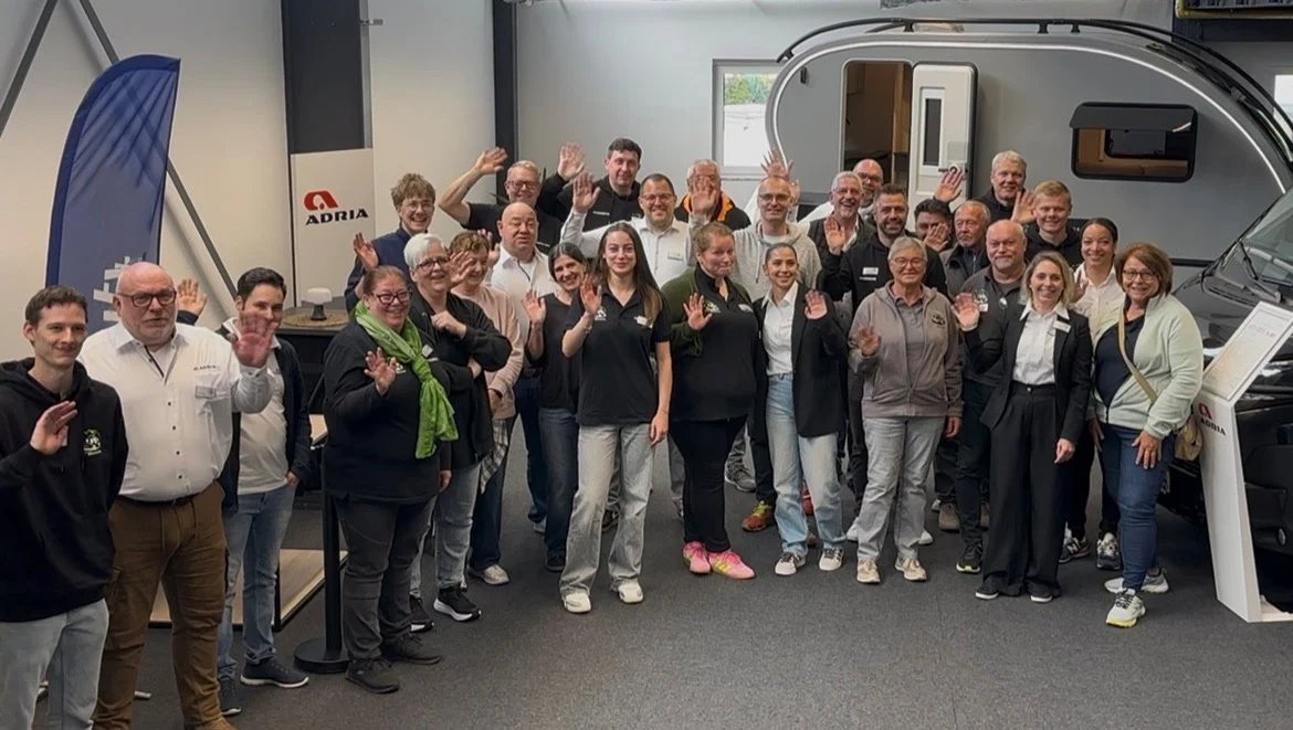 Rund dreissig Mitarbeiterinnen und Mitarbeiter stehen winkend in der Ausstellungshalle vor einem Caravan, links daneben ein Adria-Segelbanner.